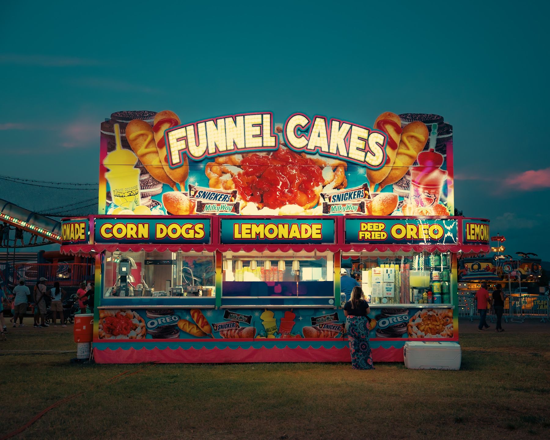 A lit fair food stand selling funnel cakes, corn dogs and lemonade
